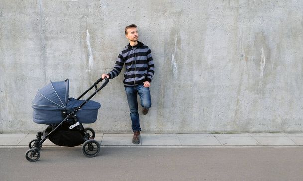 Handsome father with newborn stroller standing near gray concrete wall and looking right on copy space. Full length portrait of single parent with baby carriage. Banner. - Фото, зображення