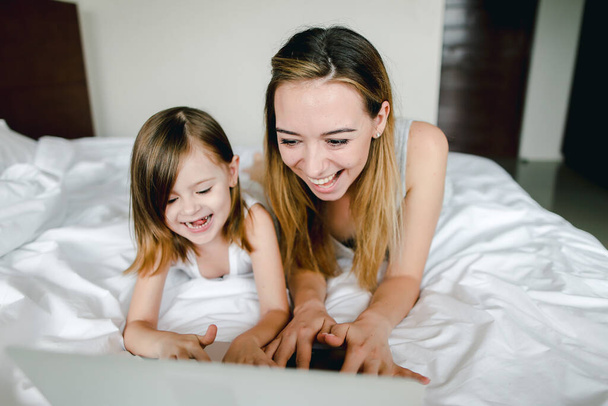 幸せな女性の人の小さな娘のラップトップを使用して、ベッドで横になっています。. - 写真・画像