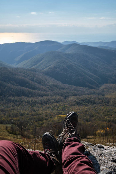Human traveler sits on top of cliff and stretches feet in boots forward, enjoying views of autumn forest and mountain peaks. First person view. Travel and explore the world. - Фото, изображение