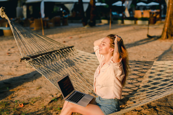 Blonde female person working with laptop and sitting on sand in wicker hammock. - Foto, imagen