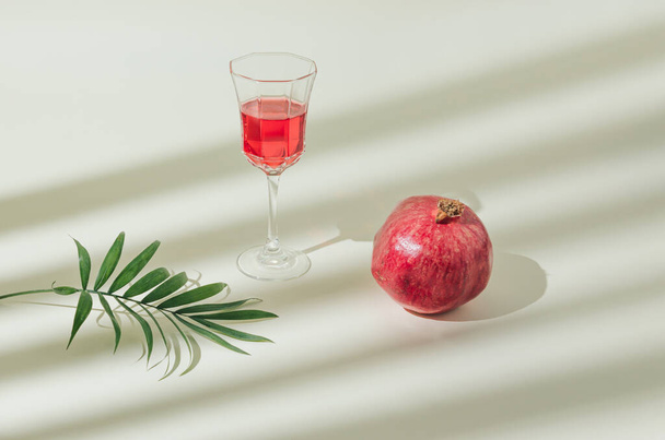 Minimal natural autumn fruit arrangement with green tropical palm leaf, red pomegranate and pomegranate juice isolated on the pastel background. Sun and shadows. Raw healthy food and beverages idea. - Photo, Image