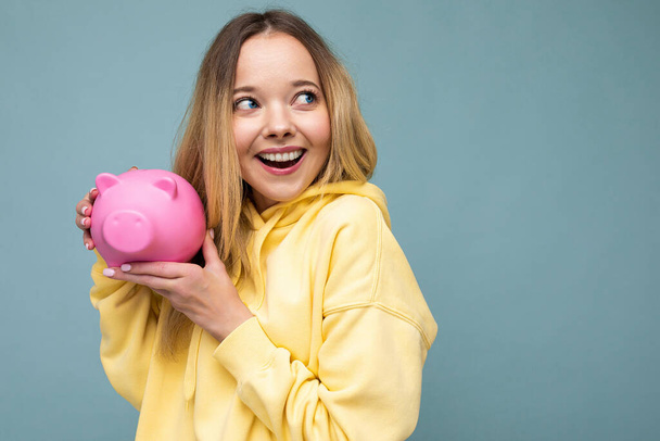 Portrait photo of happy positive smiling young beautiful attractive blonde woman with sincere emotions wearing trendy yellow hoodie isolated over blue background with copy space and holding pink pig - Zdjęcie, obraz