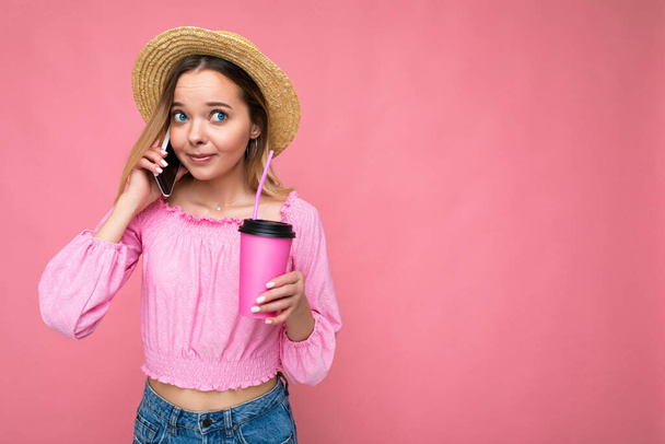 Foto de hermosa mujer rubia emocional positiva que usa blusa rosa y sombrero de paja hablando en el teléfono celular mientras bebe bebidas aisladas en el fondo rosa con espacio para copiar - Foto, imagen