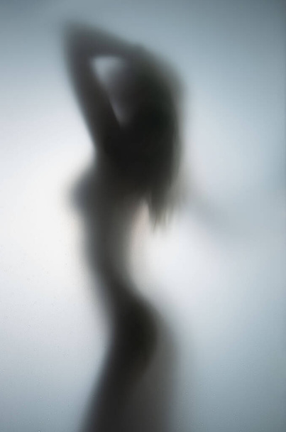 Sexy and beautiful long haired natural naked body woman stands behind a wet glass surface in bathroom. - 写真・画像