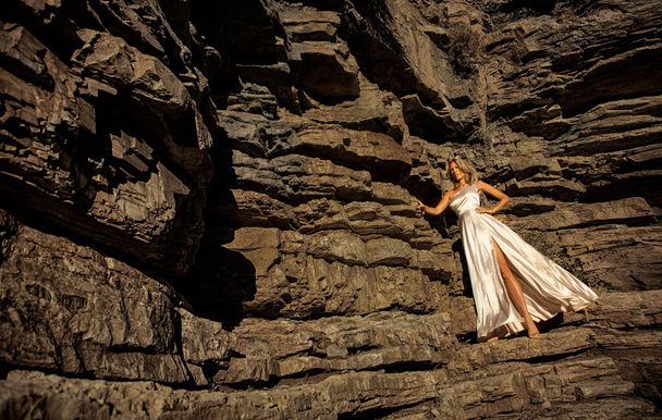 Beautiful long haired blonde milf woman in flying long beige color dress barefeet stands on rough rocks of a canyon in Rhodes, Greece  - Φωτογραφία, εικόνα