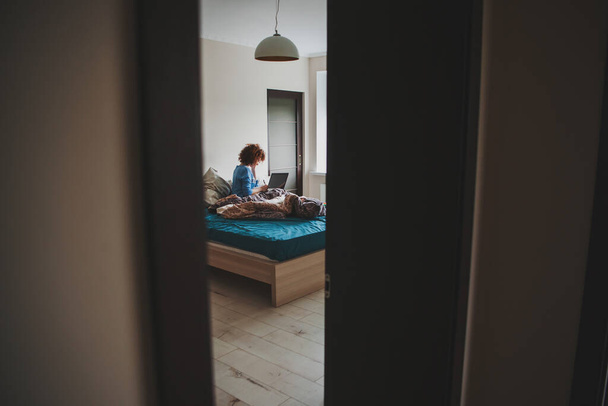 Woman with curly hair starting the morning taking online classes on laptop from the still messy bed. Online education. View from the half-open door - 写真・画像
