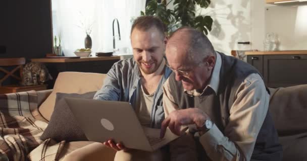 Happy smiling bearded young adult man helps senior dad teaching to use laptop, internet on sofa at home living room. - Felvétel, videó