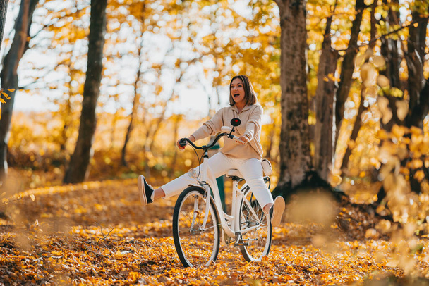Young pretty woman riding vintage white bicycle in autumn park. Lady having fun on orange nature fall background. - Foto, Imagem