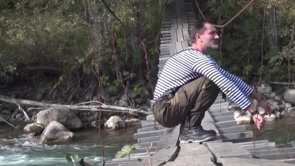 Un hombre en un chaleco se sentó en un puente colgante sobre un río de montaña. Observa detenidamente el paisaje otoñal - Imágenes, Vídeo