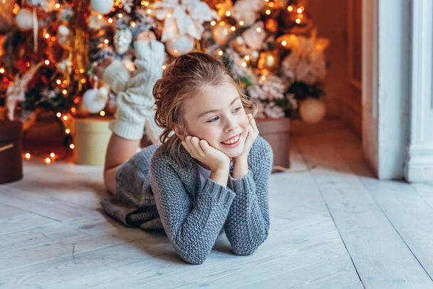 Mała dziewczynka z pudełkiem prezentów w pobliżu choinki w Wigilię w domu. Młody dzieciak w jasnej sypialni z zimową dekoracją Szczęśliwa rodzina w domu. Boże Narodzenie Nowy Rok grudzień czas na świętowanie koncepcji - Zdjęcie, obraz