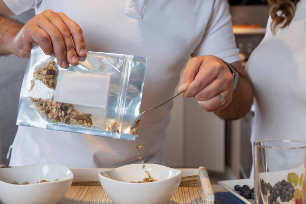 Manos de hombre vaciando granola en un tazón blanco en una cocina - Foto, imagen