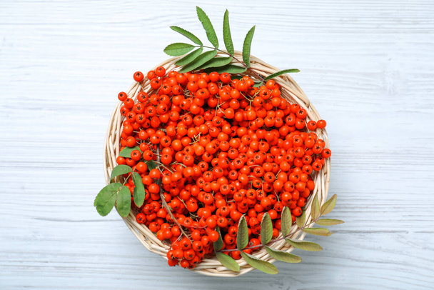 Fresh ripe rowan berries and leaves in wicker basket on white wooden table, top view - Fotó, kép