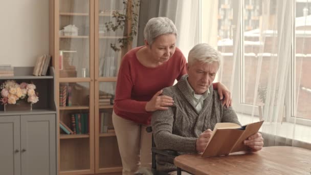 Medium long of older Caucasian man with physical disability sitting at desk by window at home, reading book, his grey-haired Asian wife hugging husband from behind, talking - Footage, Video