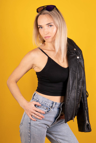 Mooie stijlvolle blonde vrouw met zwart leren jas en jeans op een gele achtergrond - Foto, afbeelding