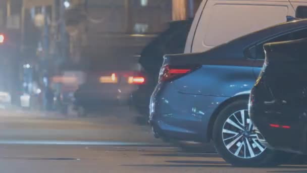 Zbliżenie zaparkowanych samochodów na poboczu drogi w nocy z rozmytym widokiem na światła ruchu pojazdów poruszających się na ulicy miasta - Materiał filmowy, wideo