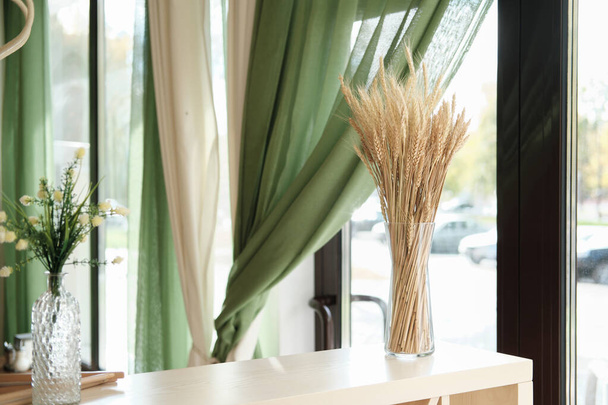 vista parcial del elegante interior de la cafetería o terraza con decoración natural. espiga de trigo en un jarrón de vidrio sobre una mesa de madera clara, cortinas verdes y flores. ventana vista a una calle. - Foto, imagen