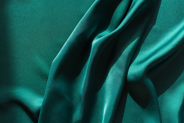 Blue-green satin fabric. Smooth silk fabric cut off on the table. Shiny soft folds in selective focus. Dark turquoise color of the sea wave. Deep rich design for covers, websites, sewing workshops. - Fotografie, Obrázek