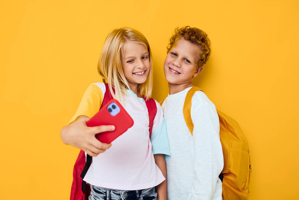 Φωτογραφία από δύο μικρά παιδιά κορίτσι με σχολική τσάντα τηλέφωνο ψυχαγωγία επικοινωνία κίτρινο φόντο - Φωτογραφία, εικόνα
