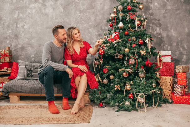 家で居心地の良い幸せな夜の若いカップルの写真,クリスマスの装飾,赤緑の色合い,ギフトボックスを持つ女性と男性.2022年大晦日。家族は楽しい - 写真・画像