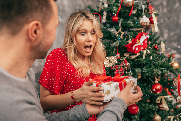 Emotions, surprise, happiness. Photo of woman. Who receives gift box from man against background of Christmas tree. Decorated in red-green colors. Happy moment - Foto, imagen