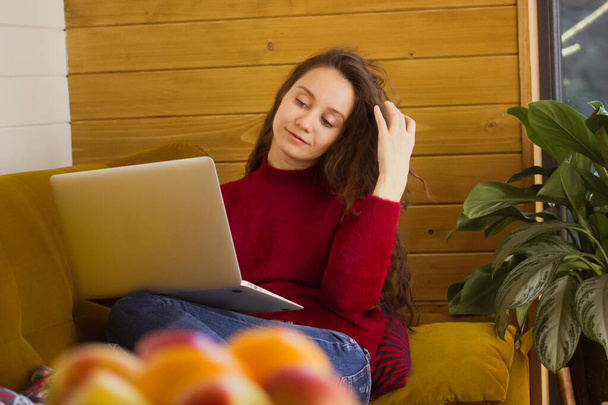 Beautiful smiling young woman, girl relaxing lying on couch in cozy home atmosphere with laptop. Online socializing, chatting, shopping, remote work. People in the interior. Stay home in fall, winter. - Photo, Image