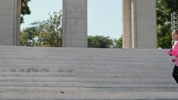 Młoda kobieta robi podział na schodach parku trzymając penny board - Materiał filmowy, wideo