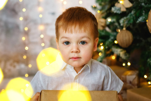 Selective focus. A boy is sitting on the floor with a gift box for a room decorated for Christmas against the background of lights of garlands. - Foto, Bild