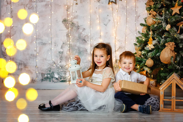 2人の子供、男の子と女の子がガーランドの明かりを背景にクリスマスのために装飾された部屋に座っています。休日のための贈り物を持つ兄弟姉妹. - 写真・画像