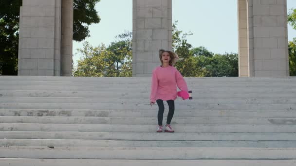 Mujer joven salta y sonríe felizmente sosteniendo penny board - Imágenes, Vídeo