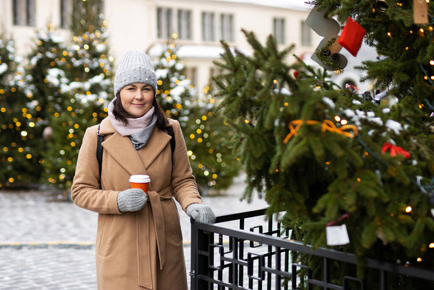 Mujer en ropa de invierno con taza de café cerca de árboles de Navidad en el casco antiguo - Foto, Imagen