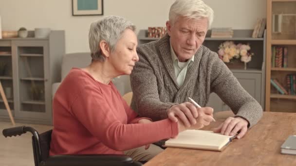 Közepes hosszúságú ősz hajú fehér férfi és idősebb ázsiai felesége tolószékben ül otthon az asztalnál, a férje segít a nőnek jegyzetfüzetben írni. - Felvétel, videó