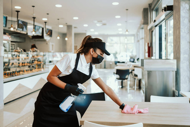 Beautiful woman working bakery or fast food restaurant. She is cleaning and disinfecting tables against Coronavirus pandemic disease. She is wearing protective face masks, gloves and face shield. - Photo, image