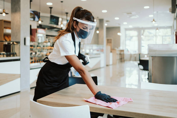 Beautiful woman working bakery or fast food restaurant. She is cleaning and disinfecting tables against Coronavirus pandemic disease. She is wearing protective face masks, gloves and face shield. - Foto, immagini