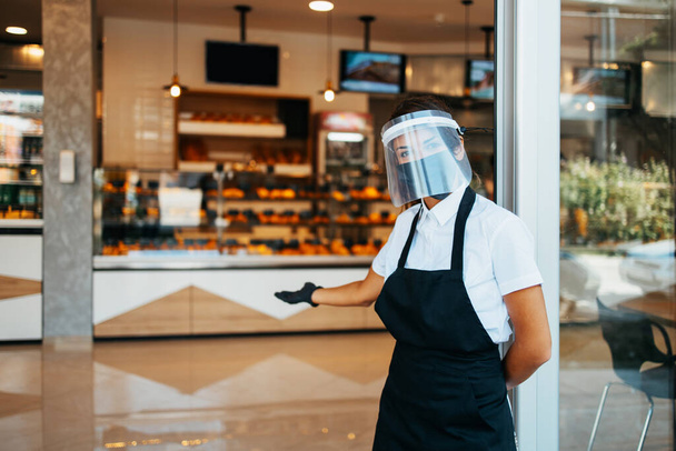 Beautiful woman with face shield and protective mask working bakery or fast food restaurant. She is standing in front shop doors and welcoming new customers. Coronavirus or Covid-19 safety measures. - Photo, image