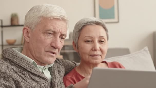 Medium close-up of senior Caucasian man hugging his cheerful Asian wife, couple using portable computer and talking, sitting on sofa in living room - Footage, Video