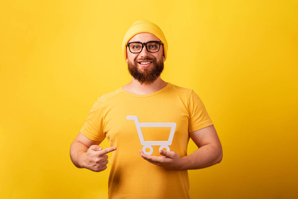 usmívající se muž ukazuje na nákupní vozík symbol v ruce přes žluté pozadí, černý pátek a velké prodejní koncepce - Fotografie, Obrázek