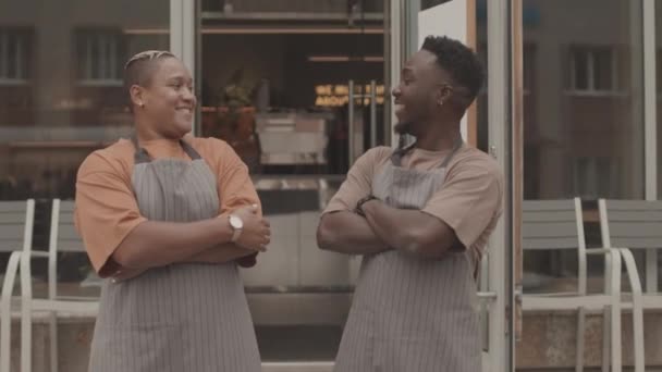Tilting up of young Black man and Biracial woman wearing aprons, standing outside restaurant, crossing arms on chests, looking and smiling on camera - Footage, Video