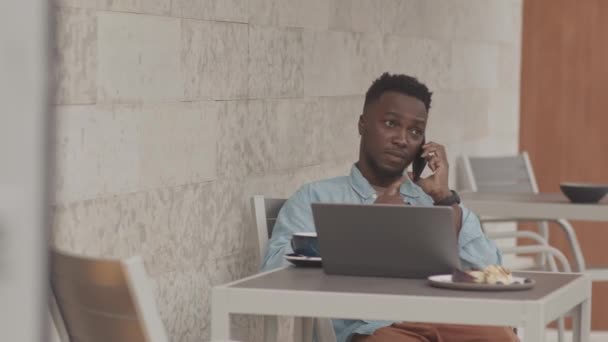 Moyen long de jeune homme noir qui est assis à la table du restaurant à l'extérieur, en utilisant un ordinateur portable au petit déjeuner, parler sur un téléphone portable, boire du café - Séquence, vidéo
