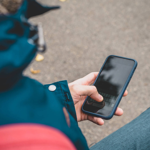 Κοντινό πλάνο του τηλεφώνου στα χέρια ενός ανθρώπου που κάθεται στην κουκέτα στο πάρκο. Άποψη από ώμο. - Φωτογραφία, εικόνα