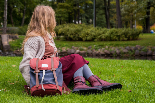 Girl in knitted jacket, burgundy skirt and leather shoes with backpack is sitting on green grass in park after school. Autumn fashion. - Photo, Image