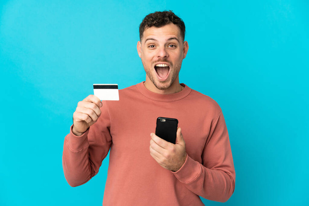 Genç, beyaz, yakışıklı bir adam mavi arka planda izole edilmiş cep telefonuyla alışveriş yapıyor ve elinde şaşırmış bir ifadeyle bir kredi kartı tutuyor. - Fotoğraf, Görsel