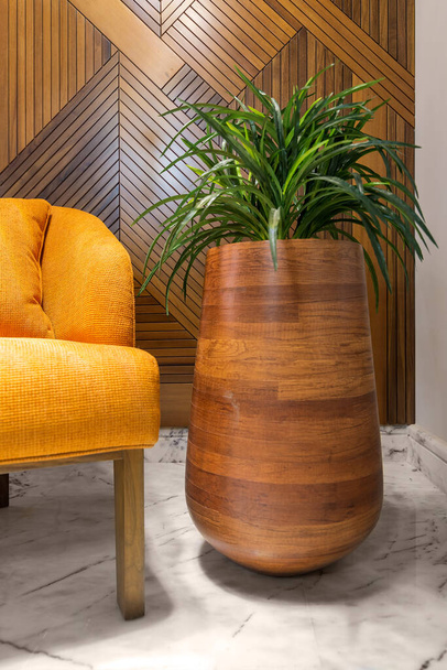 Tall rounded wooden planter with green bushes, beside orange armchair, wood cladding decorated wall, and marble floor - Photo, Image
