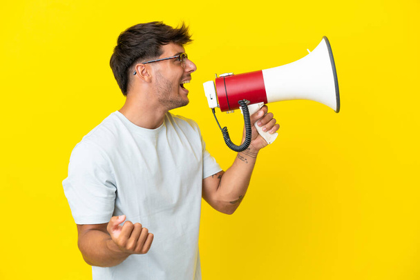 Young caucasian handsome man isolated on yellow background shouting through a megaphone to announce something in lateral position - Foto, Bild