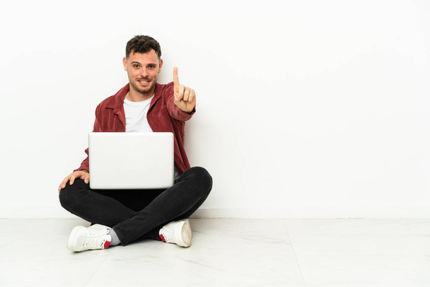 Fiatal jóképű kaukázusi férfi ül a padlón laptoppal mutatja és emeli az ujját - Fotó, kép
