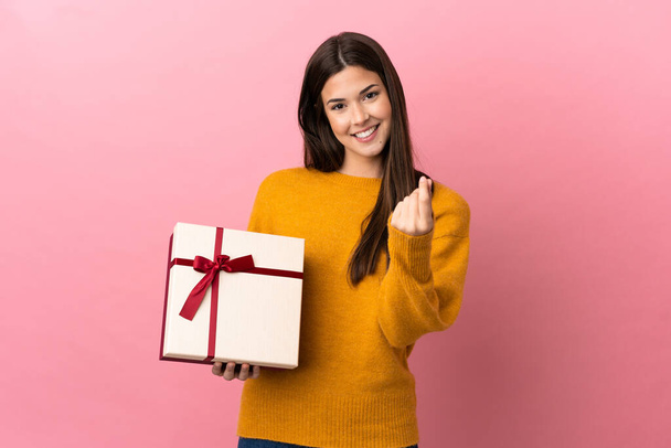 Teenager Brazilian girl holding a gift over isolated pink background making money gesture - Fotografie, Obrázek