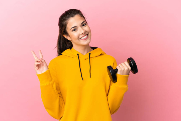 Junges Sportmädchen macht Gewichtheben auf isoliertem rosa Hintergrund lächelnd und zeigt Siegeszeichen - Foto, Bild