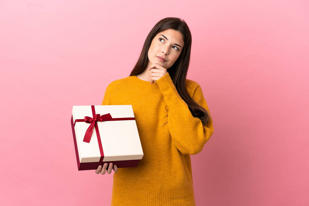 Teenager Brazilian girl holding a gift over isolated pink background and looking up - Fotoğraf, Görsel