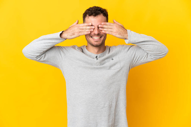 Young caucasian handsome man isolated on yellow background covering eyes by hands and smiling - Fotografie, Obrázek