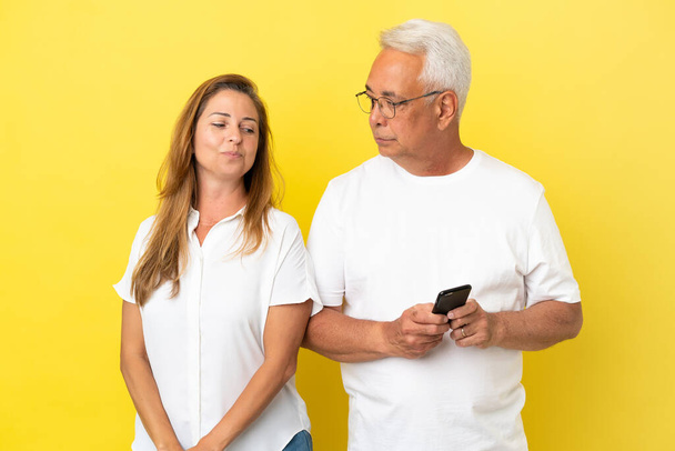 Střední věk pár izolované na žlutém pozadí čtení zprávy s mobilním - Fotografie, Obrázek