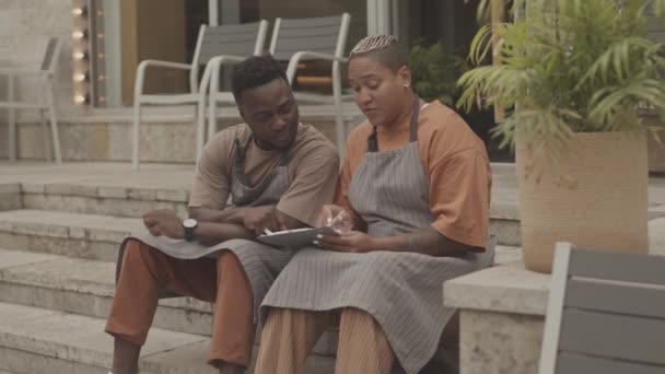 Mittlere Länge junger schwarzer Mann und Frau mit birassischen Schürzen, die auf Stufen vor dem Café sitzen und Geschäftsaufgaben besprechen - Filmmaterial, Video